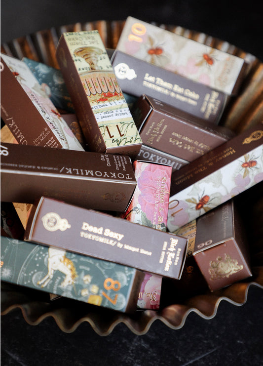 Assorted hand cream tubes by Tokyo Milk, packaged in decorated brown boxes, displayed in a gold ruffled bowl.
