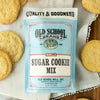Vanilla sugar cookie mix by Old School Brand, surrounded by baked sugar cookies on a white cloth.