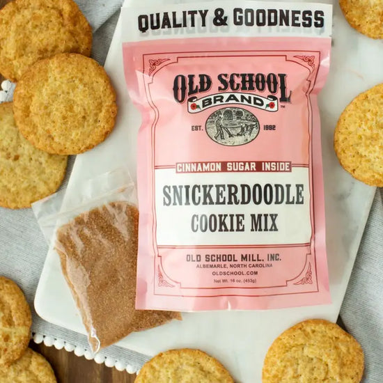 Snickerdoodle cookie mix by Old School Brand, with cinnamon sugar packet, surrounded by baked cookies on a serving board.
