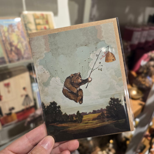 Illustration card of a bear by an unknown brand with beehive and flying bees, set on a landscape background.
