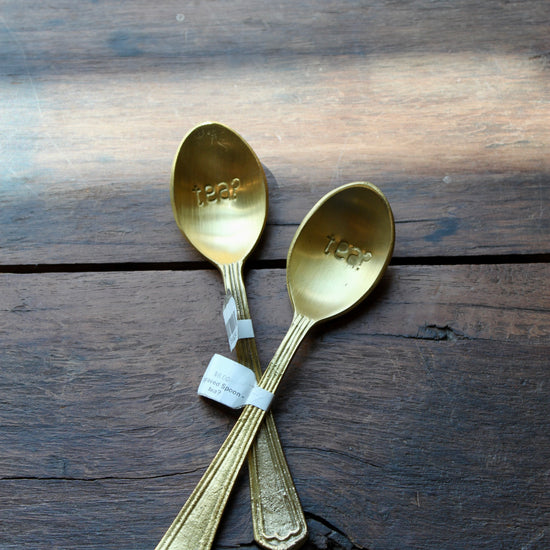 Gold-toned engraved spoons with "tea?" inscription by Creative Brands, arranged on rustic dark wood surface.