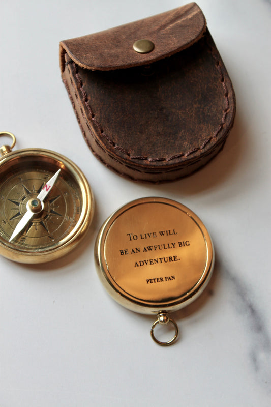 Gold compass with leather pouch, featuring a quote by Peter Pan, on a white marble surface.