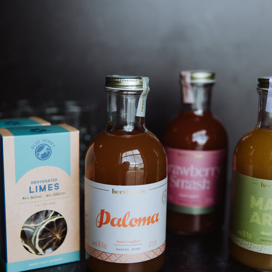 Bottles of Paloma cocktail mixer by Here's How, alongside a box of dehydrated limes, and blurry cocktail bottles in background.
