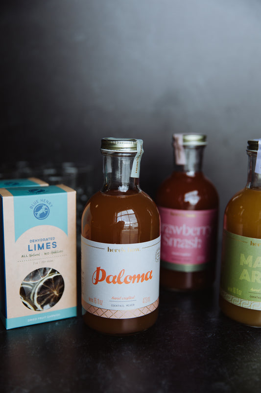 Bottles of Paloma cocktail mixer by Here's How, alongside a box of dehydrated limes, and blurry cocktail bottles in background.
