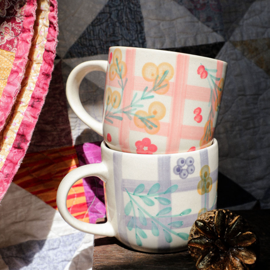 Two patterned mugs stacked on a wooden surface with a quilted fabric background.