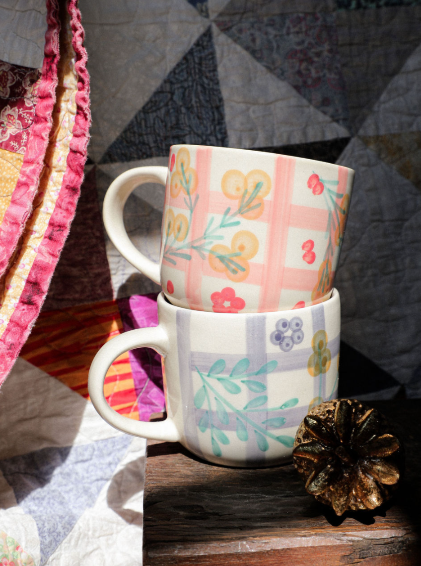 Two patterned mugs stacked on a wooden surface with a quilted fabric background.