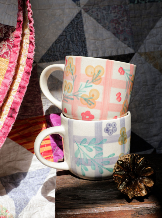 Two patterned mugs stacked on a wooden surface with a quilted fabric background.