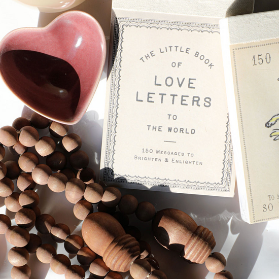 Gift book "The Little Book of Love Letters to the World" with a decorative border, and a wooden bead necklace nearby.
