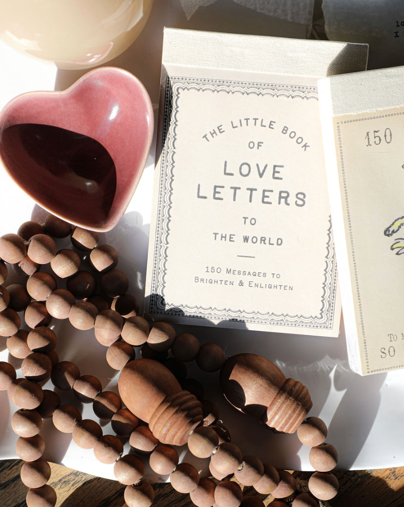 Gift book "The Little Book of Love Letters to the World" with a decorative border, and a wooden bead necklace nearby.
