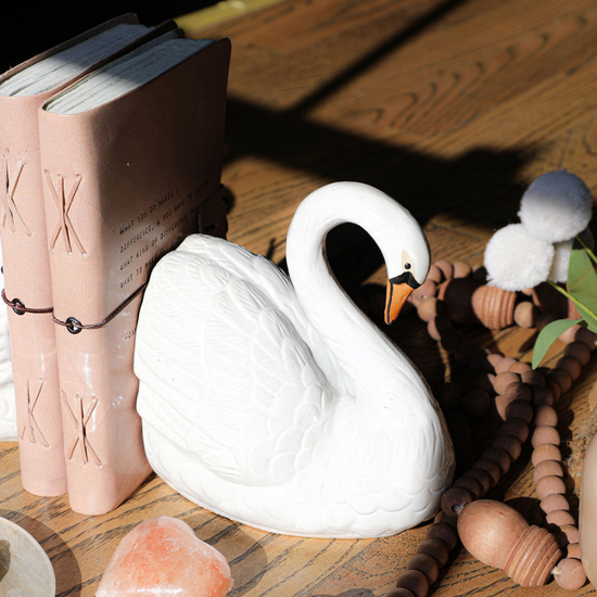Set of two stoneware swan shaped bookends with orange beaks, placed on a wooden surface among other decorations.