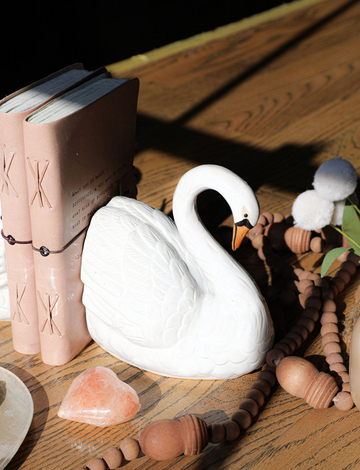 Set of two stoneware swan shaped bookends with orange beaks, placed on a wooden surface among other decorations.