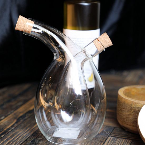 Clear glass oil and vinegar cruet with cork stoppers, on a wood table, behind an unlabeled bottle.
