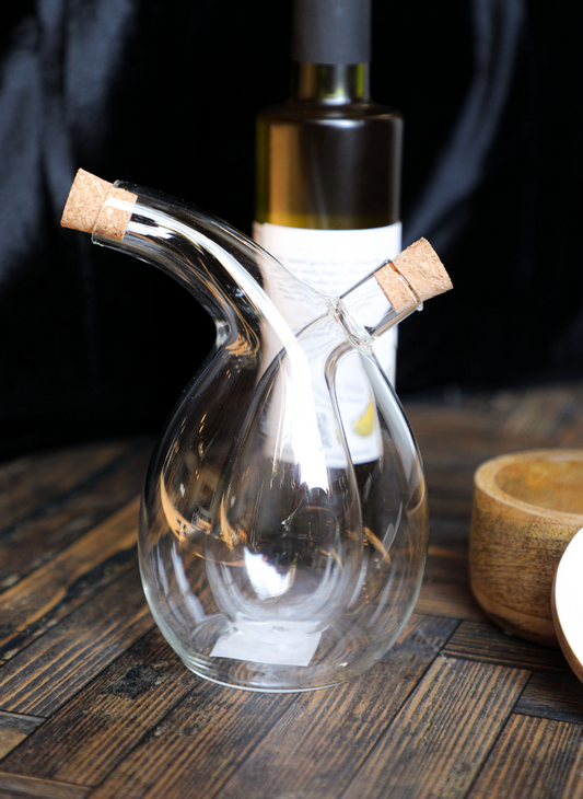 Clear glass oil and vinegar cruet with cork stoppers, on a wood table, behind an unlabeled bottle.
