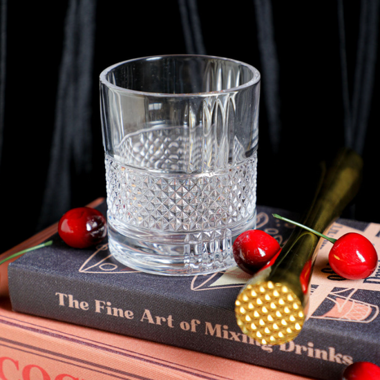 Clear Revolve Rocks Glass from Anthropologie, sitting on cocktail books with cherries and a gold cocktail muddler.