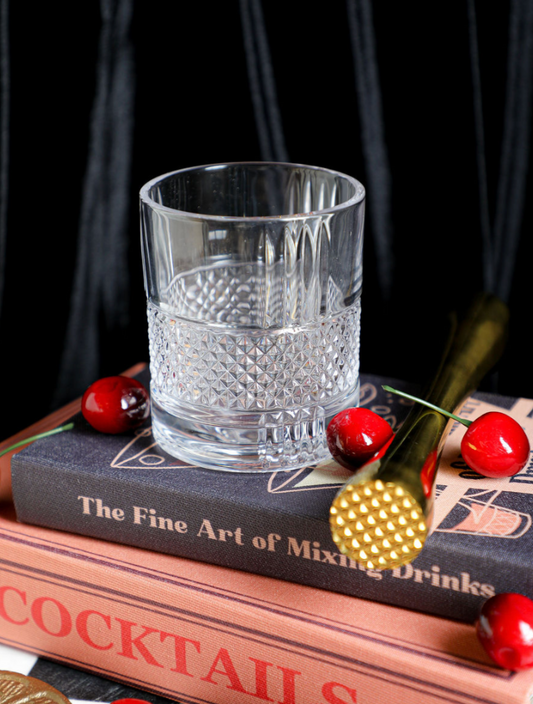Clear Revolve Rocks Glass from Anthropologie, sitting on cocktail books with cherries and a gold cocktail muddler.