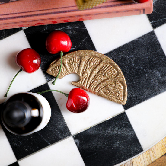 Gold metal lemon slice shaped bottle opener with textured details on a black and white checkered surface with cherries.
