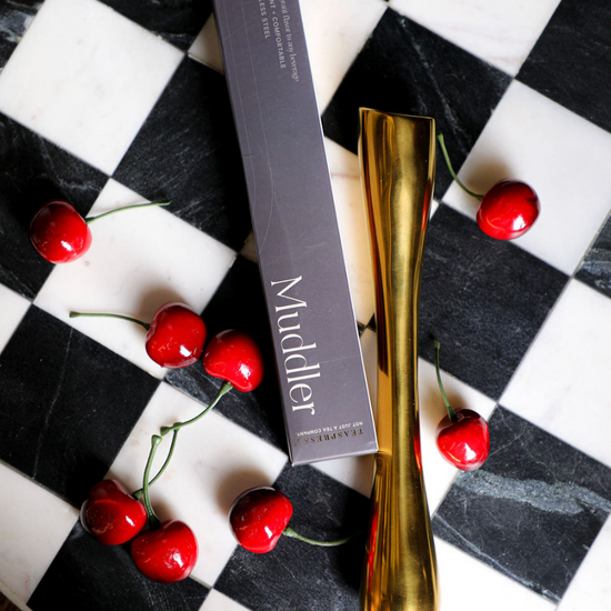 Gold muddler by TEASPRESSO set on a black and white checkerboard surface with cherries.