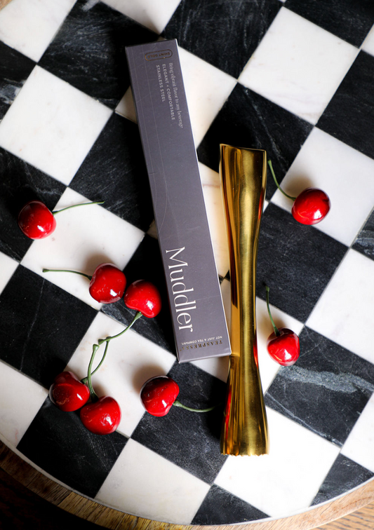 Gold muddler by TEASPRESSO set on a black and white checkerboard surface with cherries.
