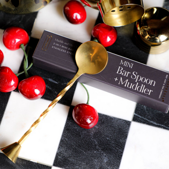 Gold mini bar spoon and muddler by Daspressa, resting on a black and white checkered surface with cherries.