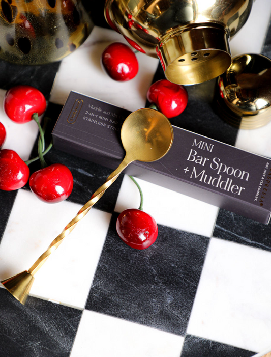 Gold mini bar spoon and muddler by Daspressa, resting on a black and white checkered surface with cherries.