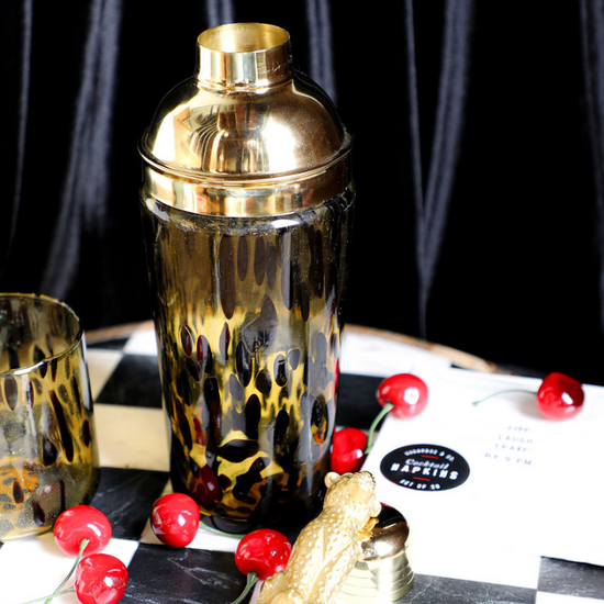 Tortoise shell glass cocktail shaker by Joanna Buchanan, with golden lid, on a checkered surface with decorative cherries.
