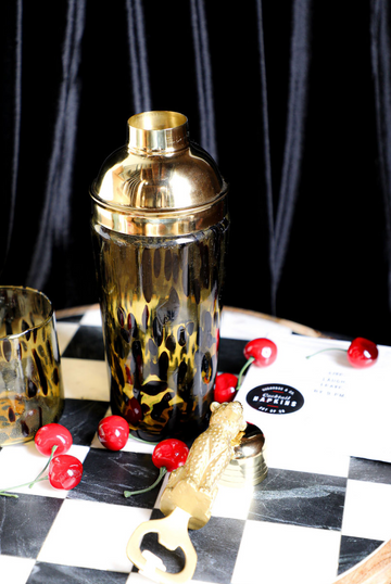 Tortoise shell glass cocktail shaker by Joanna Buchanan, with golden lid, on a checkered surface with decorative cherries.
