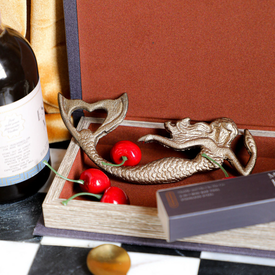 Cast iron mermaid shaped bottle opener by MHW, with a heart-shaped tail and placed among decorative cherries.