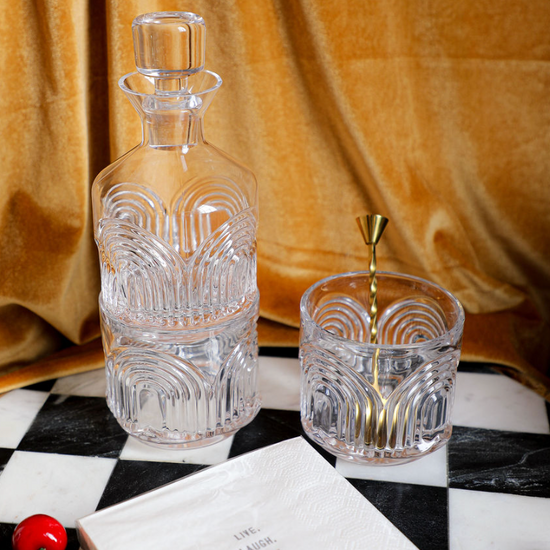 Three piece clear glass decanter set from Beau featuring a patterned design, with a matching glass and mixing spoon on a checkerboard surface.
