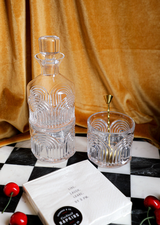 Three piece clear glass decanter set from Beau featuring a patterned design, with a matching glass and mixing spoon on a checkerboard surface.
