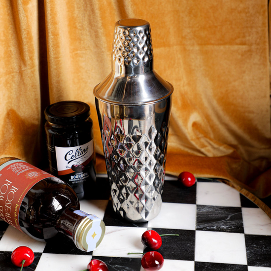 Stainless steel cocktail shaker by Crate and Barrel, on a checkered surface with dark velvet background.