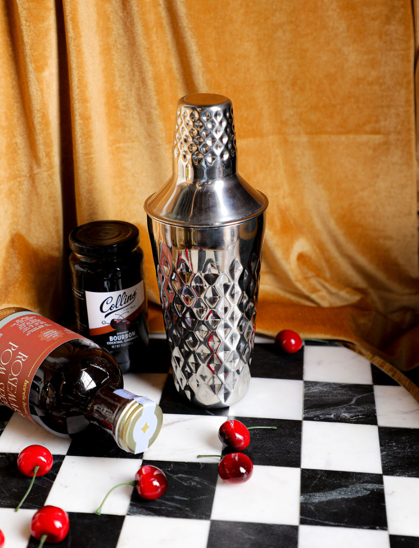 Stainless steel cocktail shaker by Crate and Barrel, on a checkered surface with dark velvet background.