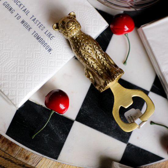 Gold leopard bottle opener from MacKenzie-Childs on a checkered marble serving board with cocktail napkins and cherries.