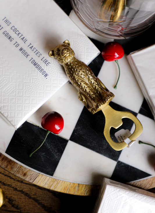 Gold leopard bottle opener from MacKenzie-Childs on a checkered marble serving board with cocktail napkins and cherries.
