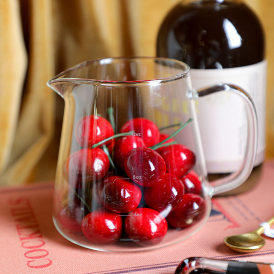 Clear glass milk frothing pitcher with ounce measurements, from an unknown brand, filled with cocktail cherries, sitting on a cocktail recipe book.
