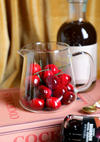 Clear glass milk frothing pitcher with ounce measurements, from an unknown brand, filled with cocktail cherries, sitting on a cocktail recipe book.
