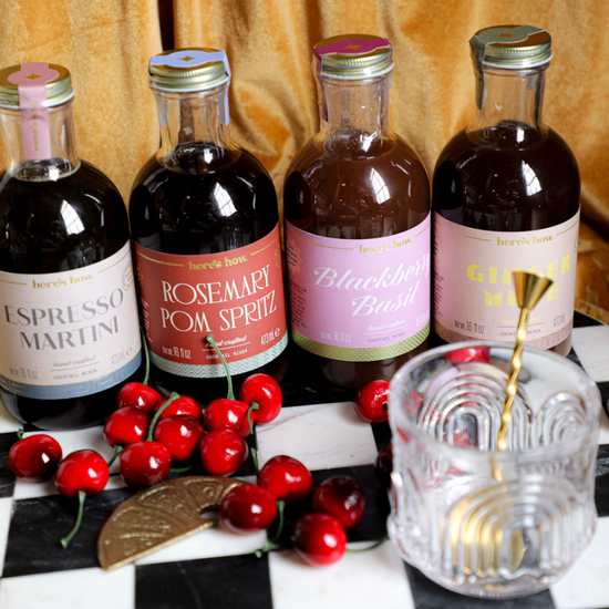 Assorted cocktail mixers by Here's How, displayed with cherries, a bar spoon, and a glass on a checkered tray.
