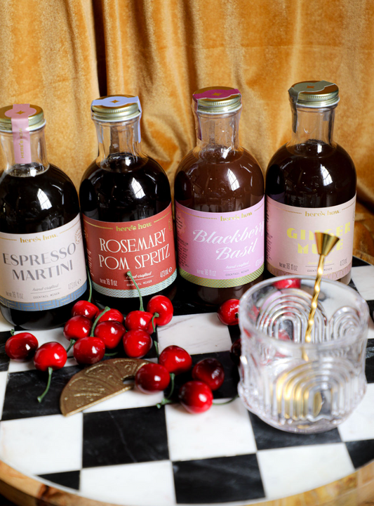 Assorted cocktail mixers by Here's How, displayed with cherries, a bar spoon, and a glass on a checkered tray.