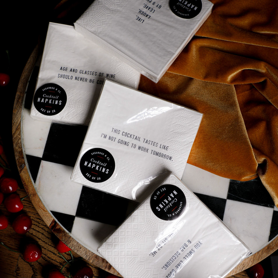 White cocktail napkins by Sugarboo & Co. with various drinking-themed sayings displayed on a checkered serving tray.