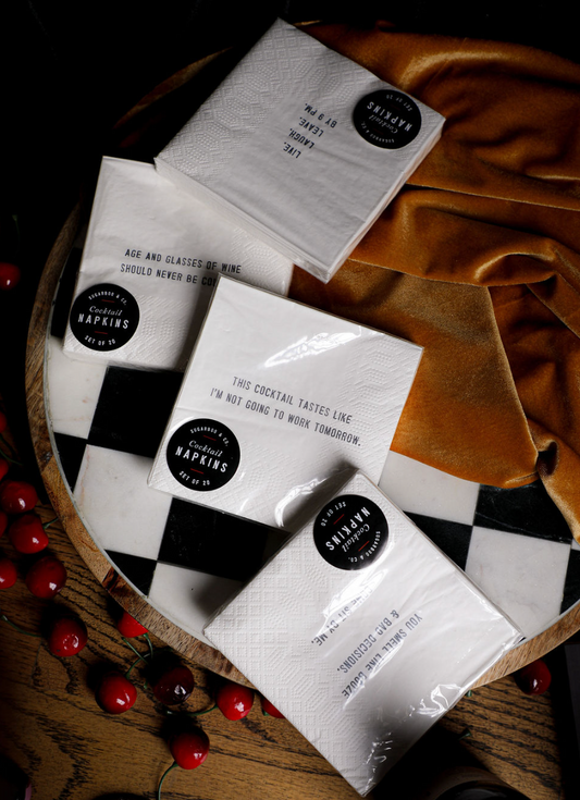 White cocktail napkins by Sugarboo & Co. with various drinking-themed sayings displayed on a checkered serving tray.