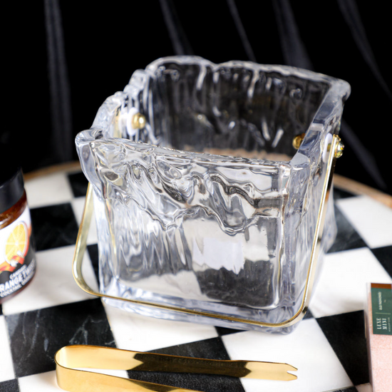 Textured glass ice bucket with tongs, with gold accents, on a checkered surface.