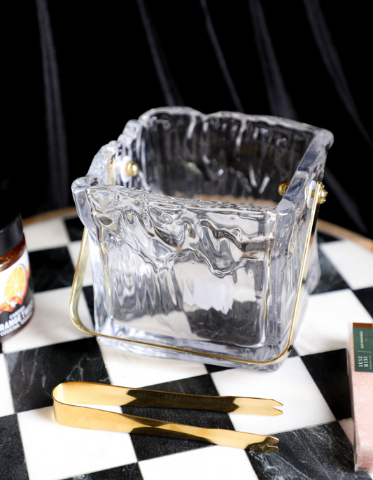 Textured glass ice bucket with tongs, with gold accents, on a checkered surface.