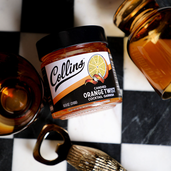 Candied orange twist in syrup by Collins, in a glass jar beside a brass bottle opener on a black and white checkered surface.