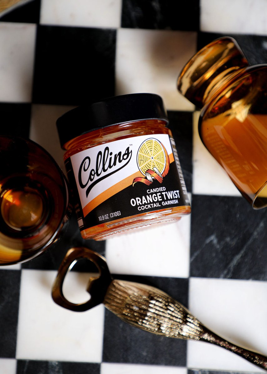 Candied orange twist in syrup by Collins, in a glass jar beside a brass bottle opener on a black and white checkered surface.