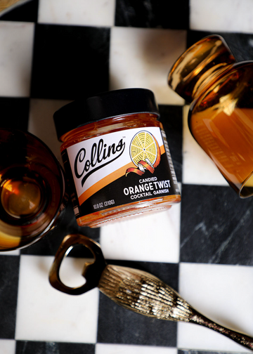 Candied orange twist in syrup by Collins, in a glass jar beside a brass bottle opener on a black and white checkered surface.
