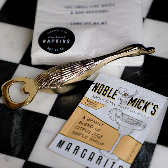 Gold duck bottle opener with textured detailing, alongside cocktail napkins and a margarita mix packet.