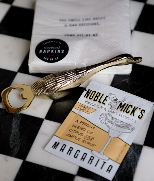 Gold duck bottle opener with textured detailing, alongside cocktail napkins and a margarita mix packet.