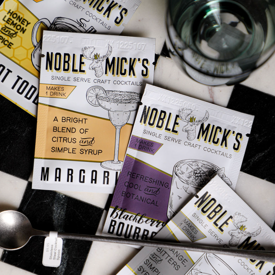 Single serve cocktail packets by Noble Mick's, with cocktail glass and spoon on a checkered surface.
