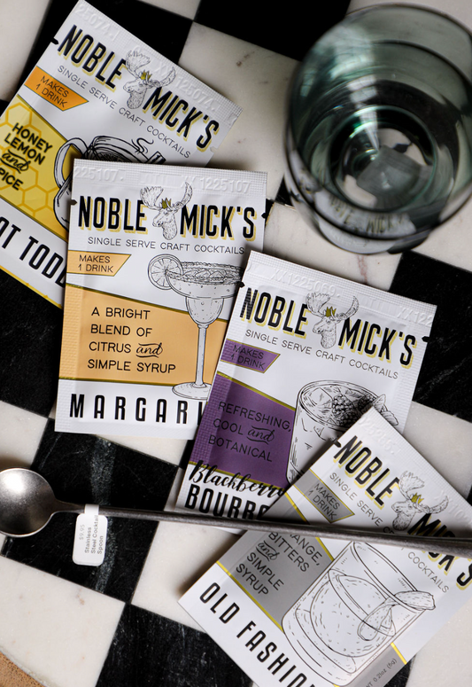 Single serve cocktail packets by Noble Mick's, with cocktail glass and spoon on a checkered surface.
