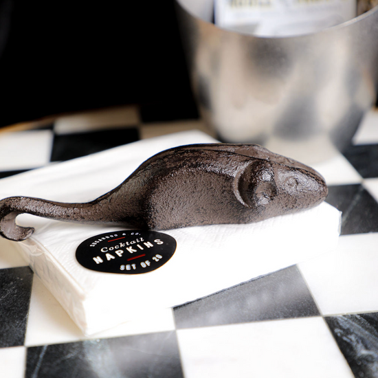 Cast iron mouse-shaped bottle opener by Sugarboo & Co., sitting on cocktail napkins on a checkered surface.