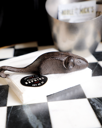 Cast iron mouse-shaped bottle opener by Sugarboo & Co., sitting on cocktail napkins on a checkered surface.